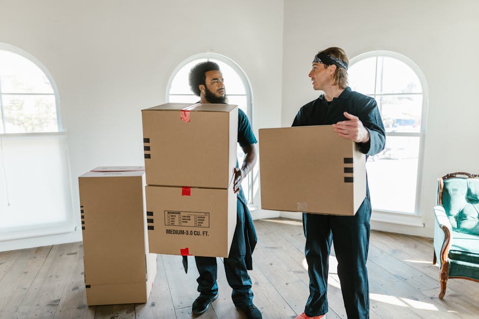 A young girl lying inside a large cardboard box during a house relocation, with her face visible and smiling up at a person outside the box. The box is on a flat surface and is partially filled with p