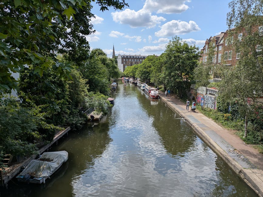 A narrow canal flanked by lush green trees and residential buildings on the right side, with a paved walkway alongside the water. Several cars are parked along the walkway, and a few pedestrians are walking. A boat covered with a protective tarp is moored to the canal bank on the left. The sky is partly cloudy with patches of blue visible, and the scene suggests a peaceful urban environment during daytime. This setting is typical for house removals and furniture transport in the Clapham area, with the canal providing a scenic backdrop for home relocation activities.