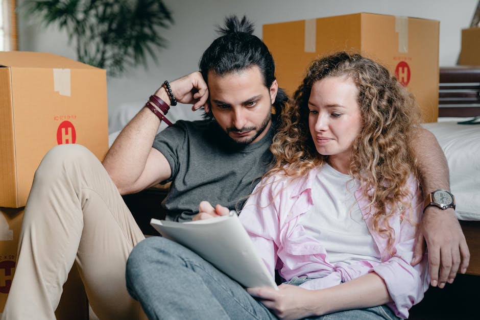 A couple sits on the floor of a room filled with cardboard moving boxes, some sealed with packing tape labeled with a red 'H,' indicating packing in preparation for house removal. The man, with dark hair tied in a bun and wearing a grey T-shirt, has his arm around the woman and appears to be thinking or resting during the packing process. The woman, with curly blonde hair, wears a light-colored shirt and holds a clipboard or folder, possibly reviewing a packing checklist. Behind them, additional large boxes are stacked near a doorway, suggesting a move-in or move-out phase as part of a home relocation or furniture transport. The scene is well-lit, capturing the interior environment where packing materials such as cardboard boxes are visible, and the individuals are engaged in the relocation planning process, supported by the packing and moving services of Clapham Man and Van.