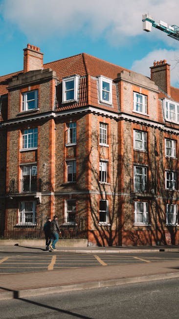 The image depicts the exterior of a multi-storey residential building with red brick walls and several windows, some of which have white frames and are open. The building has a sloped red tile roof with dormer windows, and tall brick chimneys extend from the roof. In front of the building, on the sidewalk, two individuals are walking away from the camera, dressed in casual clothing. Shadows of leafless trees are cast onto the building's facade. A section of a loading area or pavement is visible at the base of the building, bordered by yellow markings indicating parking or loading zones. The sky is mostly clear with some clouds, and part of a blue construction crane is visible in the upper right corner. The scene suggests a typical urban residential area, with the presence of [COMPANY_NAME] and the context of home relocation or furniture transport implied by the surroundings.
