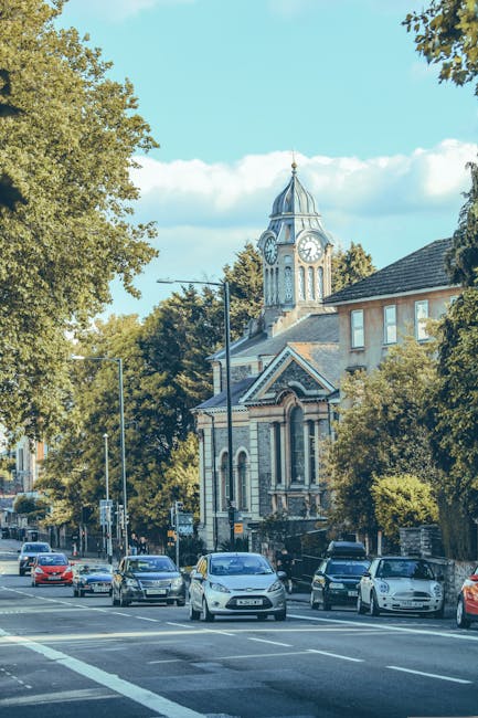 A busy street scene in Clapham featuring a row of parked cars along the curb, with a variety of vehicles including sedans and hatchbacks. In the background, there is a historic building with a promine